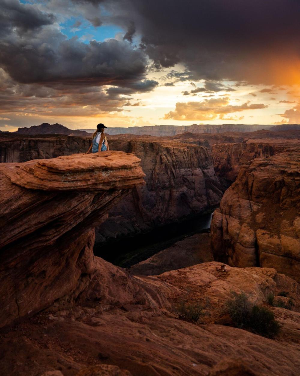 Person on rocky cliff overlooking canyon at sunset with dramatic clouds.