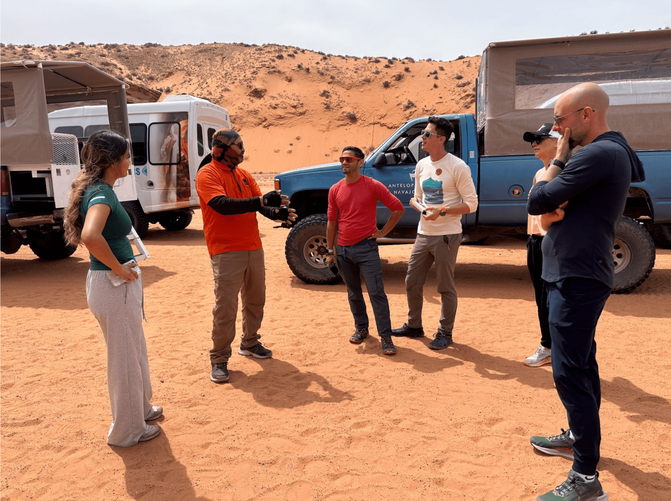Group of six people talking near off-road vehicles on a desert landscape.